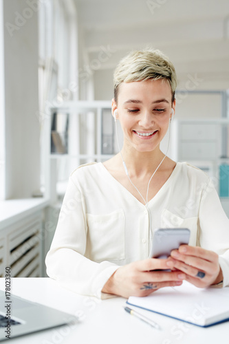 Positive young businesswoman choosing her favorite tracks from playlist in smartphone by workplace