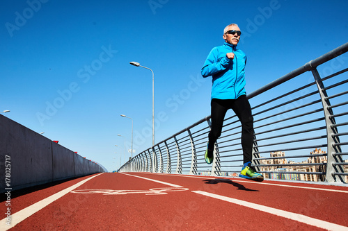Low angle view of concentrated senior sportsman jogging outdoors and enjoying sunny warm day, full length portrait
