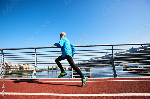 Profile view of sporty senior man running on modern bridge while preparing for marathon, picturesque cityscape on background