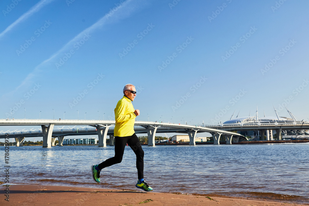 Profile view of enthusiastic aged sportsman enjoying fresh air while running along lakeside, cloudless blue sky and facade of modern building on background