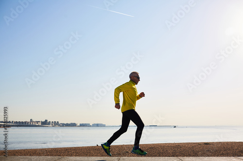 Profile view of elderly man in sportswear keeping healthy lifestyle jogging along seashore and enjoying fresh air, cloudless blue sky on background