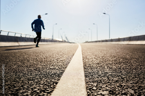 Close-up shot of modern asphalt road bridge, unrecognizable sporty man jogging while having outdoor workout, cloudless blue sky