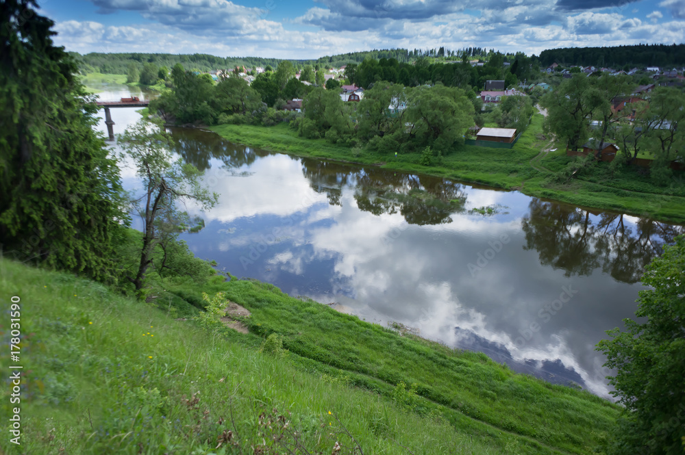 Fototapeta premium View of the blue river from a high green hill. The beauty of nature. Blue sky with clouds and green summer around.