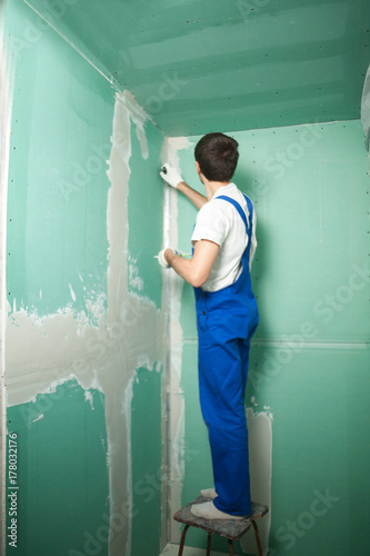 Portrait of young pretty man on construction site: male worker plastering walls with spatula while remodeling office
