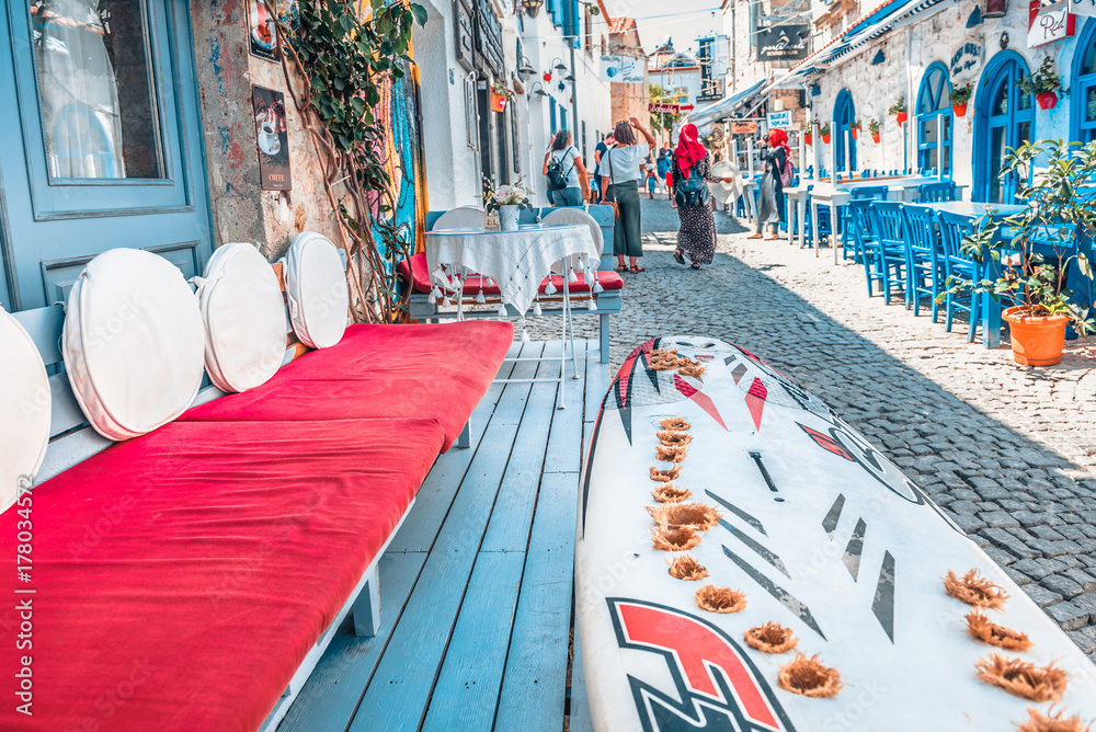 Obraz premium Unidentified People walk at a street with cafe and restaurant with tables and chairs around at Alacati Town,a popular destination for traveling and vacation in Izmir,Turkey.26 August 2017.