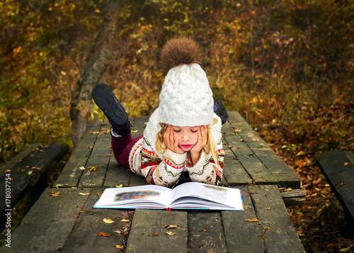 Девочка в теплой белой вязаной шапке сидит на лавочке в парке и читает книгу. Девочка с книгой.
