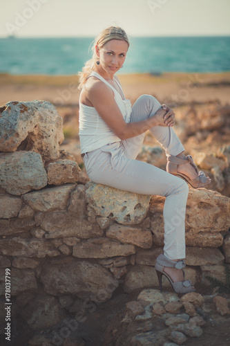 Amazing lady blond woman in light white stylish clothes sexy posing on sea side beach air.Sparkler girl looking to camera on ancient city stones blue ocean on background. photo