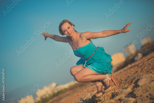 Seductive blond lady woman with sexy naked legs shoulders and arms wearing light blue open dress posing enjoying vacation time on sea side ocean beach alone lonely.