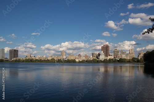 Urban city landscape of buildings and skyscrapers behind the lake or river and trees, with reflection of buildings in water and clouds on blue sky