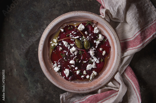 Vegetarian beetroot carpaccio salad with feta cheese, thyme, olive oil and capers in terracotta plate on textile napkin over old metal background. Top view, space. Dark rustic style, healthy eating