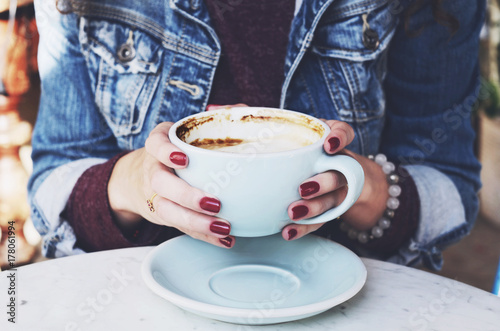 Woman in jeans jacket holding cup of cappuccino in her hands with red nails