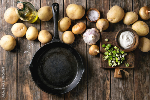 Ingredients for cooking dinner. Raw whole washed organic potatoes, onion, garlic, salt, olive oil, cast-iron pan, spring onion and cream-fresh over old wooden plank background. Top view with space