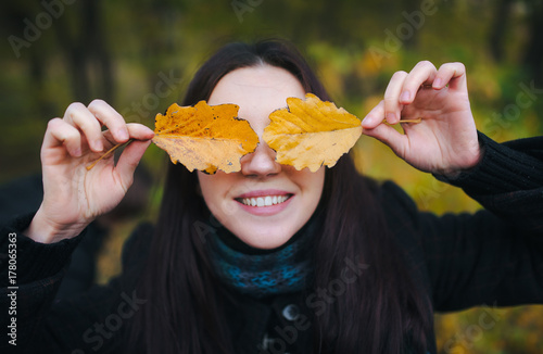 Young lovely woman covered her eyes with yellow autumn leaves and cheerfully smiles. walking in the autumn park and fun with yellow autumn leaves.