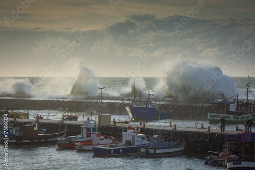 Fishing boats shelter in the harbor as huge storm waves break on the harbor wall.  Kalk Bay fishing harbor, Cape Town, South Africa