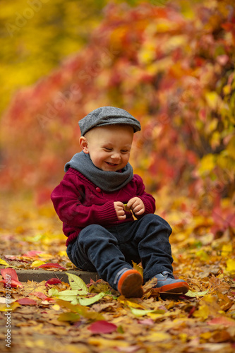Little smiling child boy sits in park and holds yellow leaf in his hands on colorful autumn background.