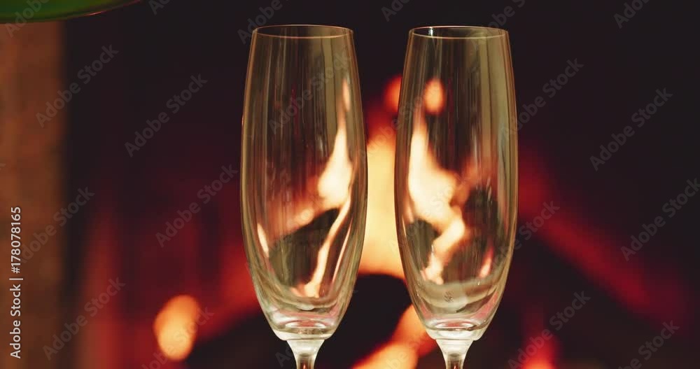Pouring two glasses of sparkling champagne in front of warm fireplace. Slow motion. Zoom out. Dolly shot. 4k graded from RAW. 