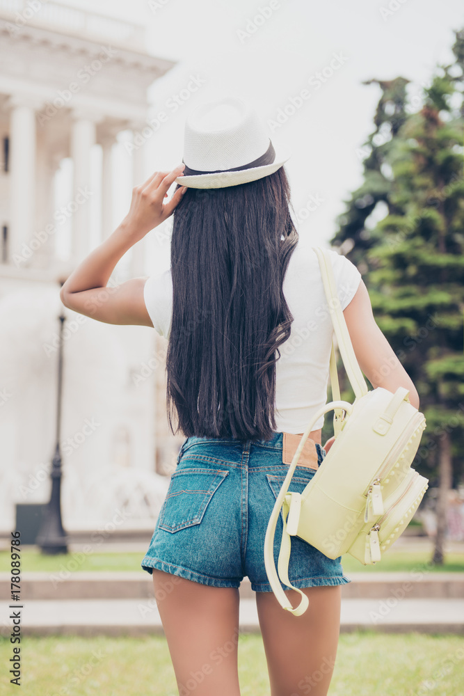 Back shot of mixed-race lady`s butt in tight short denim shorts on town`s  background. Nice smooth bronze skin, beige cap headwear, long hair, yellow  rucksack. She is on the stroll outside Stock