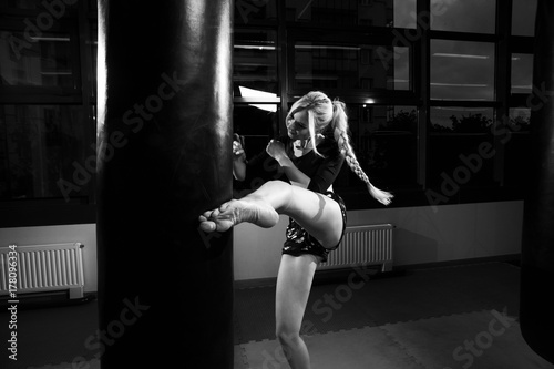Gorgeous female fighter with blonde hair pulled back in long braid, dressed in black crop top and camouflage shorts, hitting punching bag with her leg. Attractive young woman training in sports club.