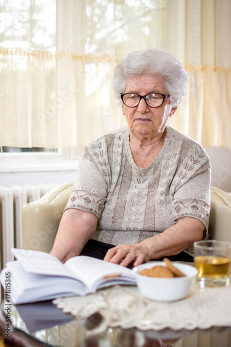 Old woman sitting on armchair and browsing through the open book at home.