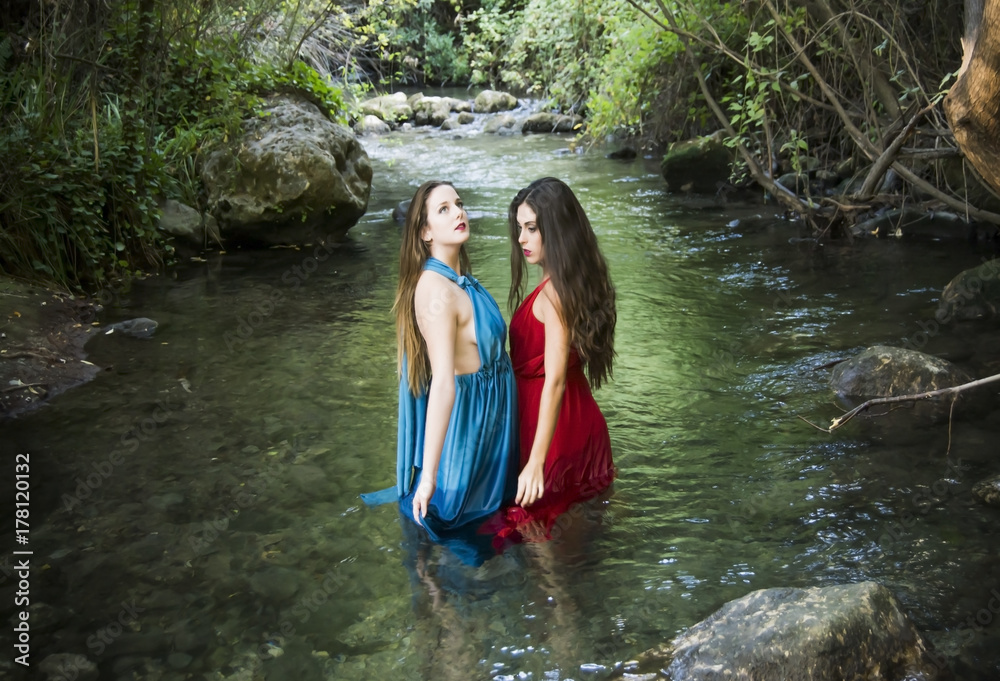 Fototapeta premium Two beautiful women on the bank of a mountain river with evening dresses