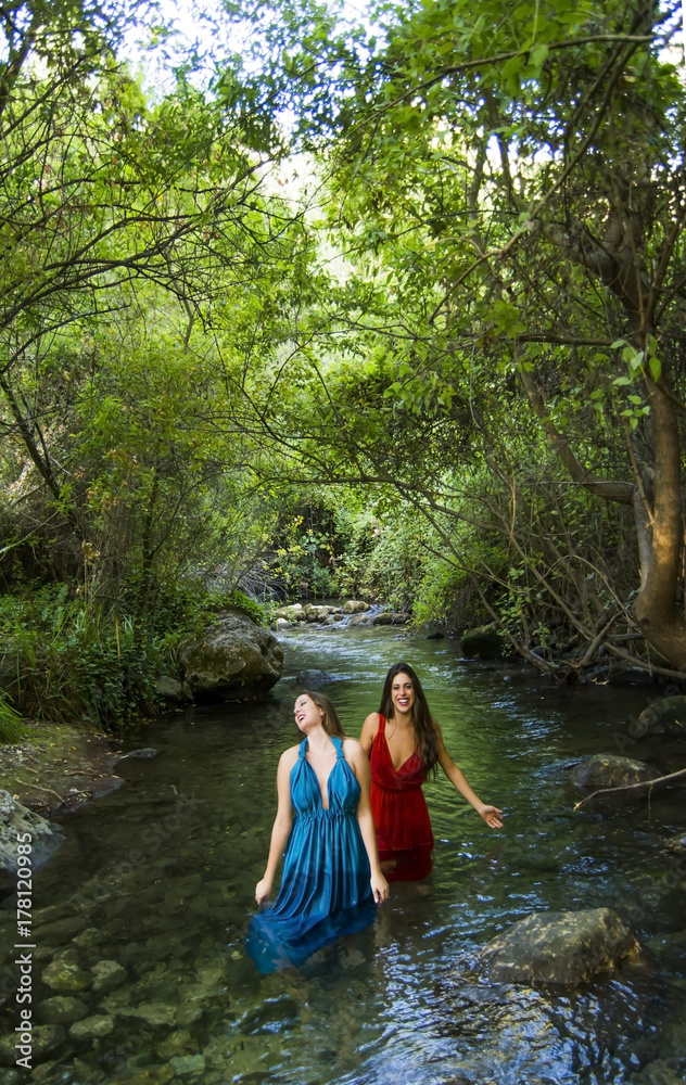 Fototapeta premium Two beautiful women on the bank of a mountain river with evening dresses