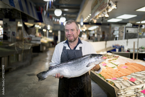 Male fishmonger wearing an apron holding large and whole salmon fish in front of display counter early in the morning on a market in England, UK. photo