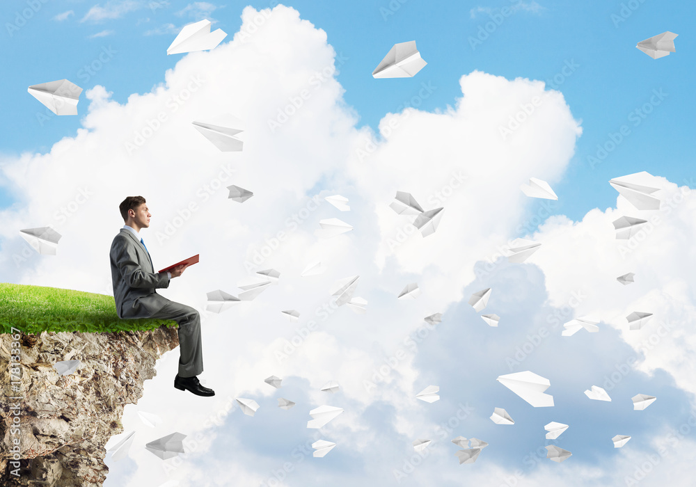 Man on edge reading book and paper planes flying in air