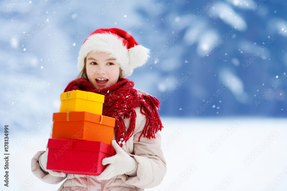 Adorable little girl wearing Santa hat holding a pile of Christmas gifts on beautiful winter day Adorable little girl wearing Santa hat holding a pile of Christmas gifts on beautiful winter day