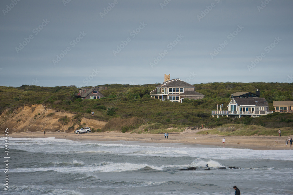 Wide exterior view of beachfront luxury vacation homes along the ocean coastline on a summer day. Rough surf waves splash along the sand