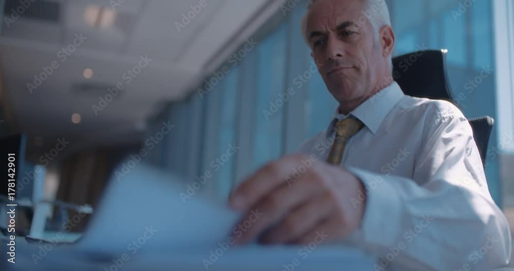 Shot of senior business man sitting at his desk and working. Business manager reading a document and working on computers in office.