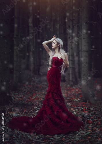 Beautiful woman with long white hair posing in a luxurious red dress with a long train standing in a autumn pine forest. Creative colors and Artistic processing.