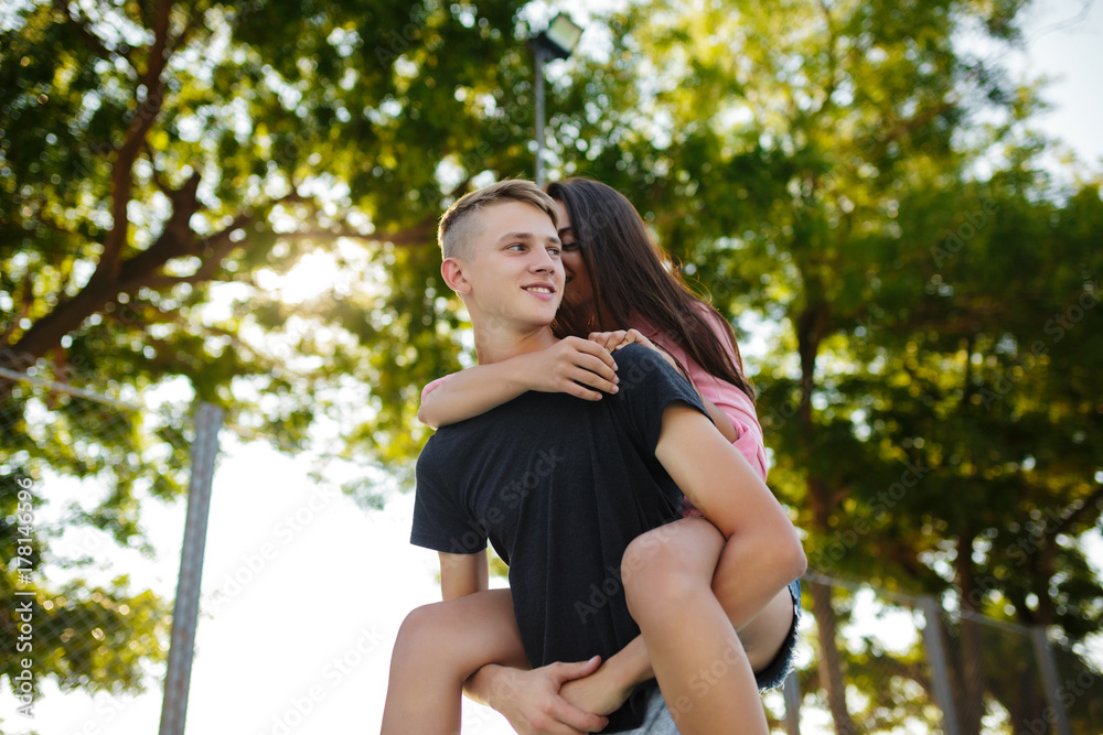 Portrait of young smiling boy holding pretty girl on his back and happily looking aside while spending time in park Portrait of young smiling boy holding pretty girl on his back and happily looking aside while spending time in park