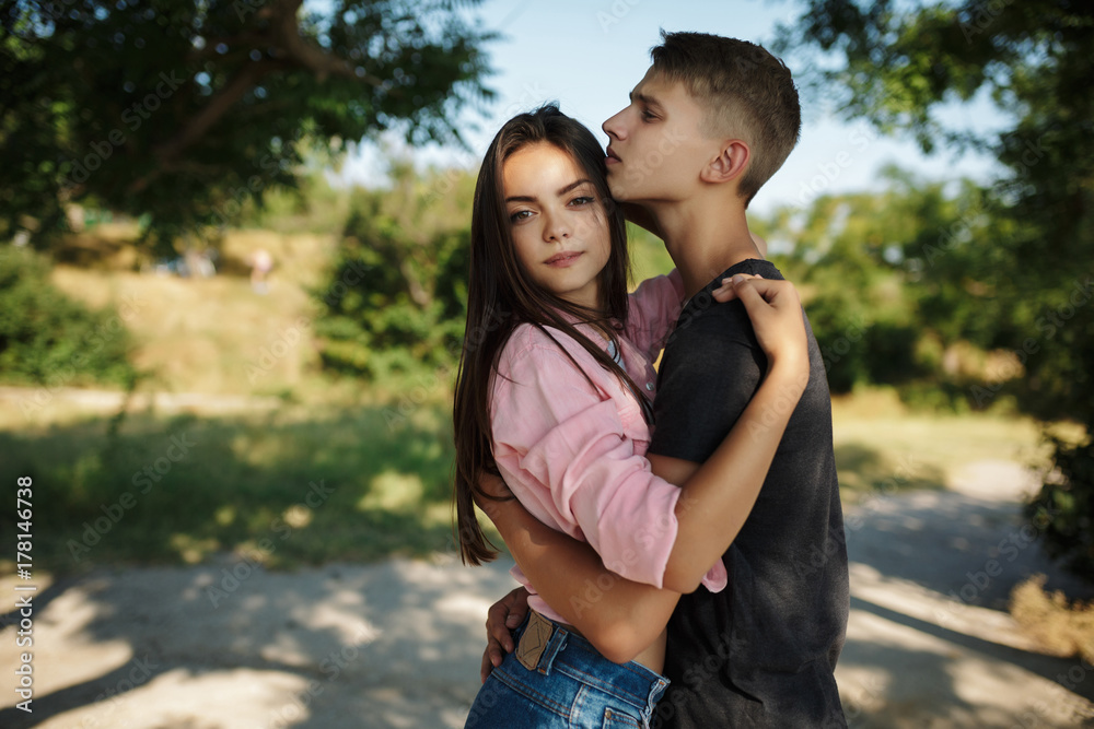 Fototapeta premium Portrait of beautiful girl thoughtfully looking in camera while standing and embracing with cool boy in park