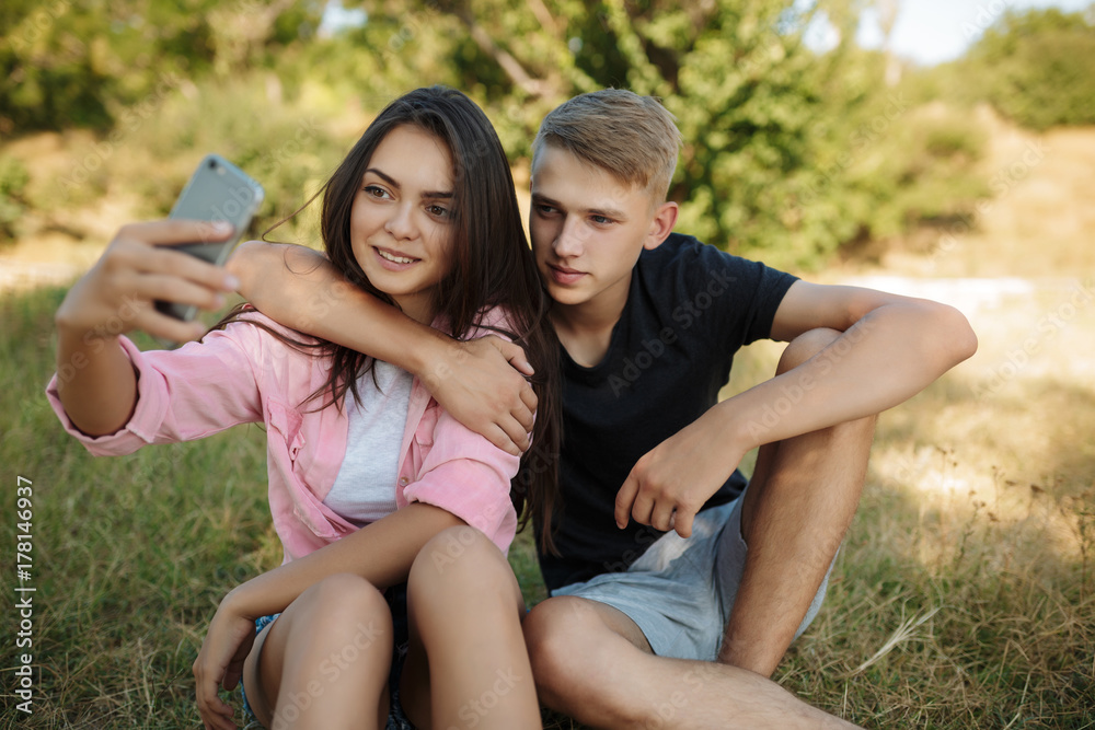 Fototapeta premium Portrait of young pretty couple sitting on lawn in park and making selfie together. Cute couple taking photos on cellphone frontal camera