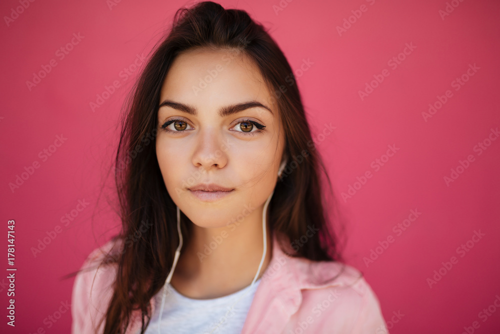 Close up photo of young thoughtful girl with dark hair standing and listening music in earphones while dreamily looking in camera on pink background isolated Close up photo of young thoughtful girl with dark hair standing and listening music in earphones while dreamily looking in camera on pink background isolated