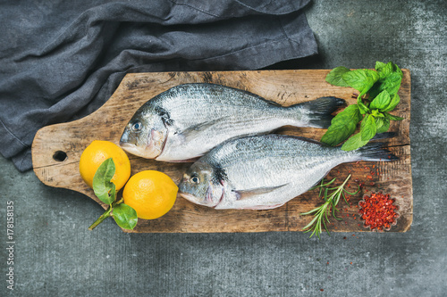 Fresh uncooked sea bream or dorado fish with lemon, herbs and spices on rustic wooden board over grey concrete background, top view. Healthy, dieting, clean eating concept