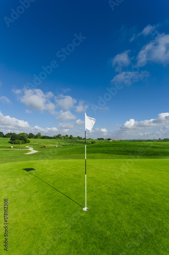 View of a green golf course, hole and flag on a bright sunny day. Sport, relax, recreation and leisure concept. Summer landscape with sunbeams