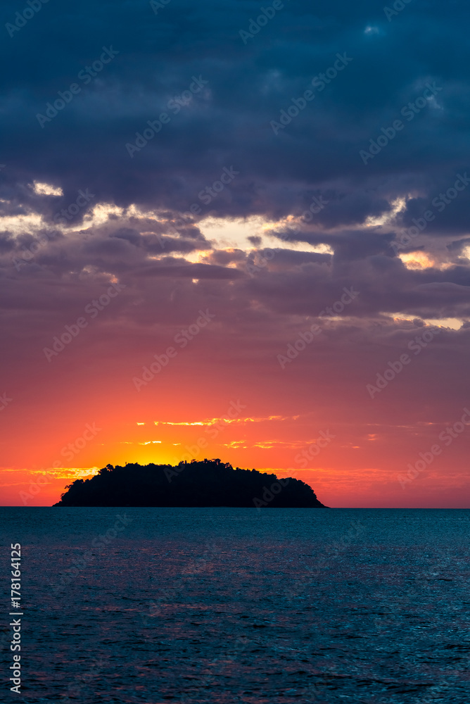 silhouette of the island at red and orange and deep blue sunset illuminated from behind by the sun on the sea with beautiful glowing clouds