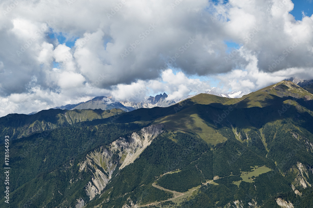A view of the mountains with forests and meadows and snow-capped peaks under the clouds