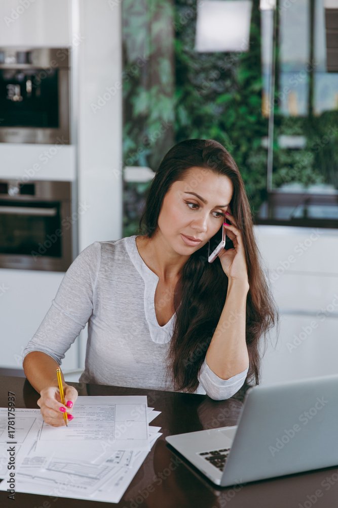 Attractive young modern business woman talking on mobile phone and working with documents and laptop in the kitchen at home Attractive young modern business woman talking on mobile phone and working with documents and laptop in the kitchen at home