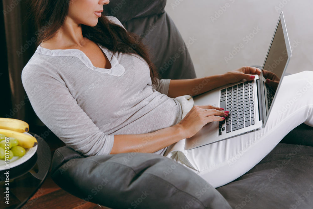 Naklejka premium Beautiful young woman at home sitting on modern chair in front of window, relaxing in her living room and working with laptop