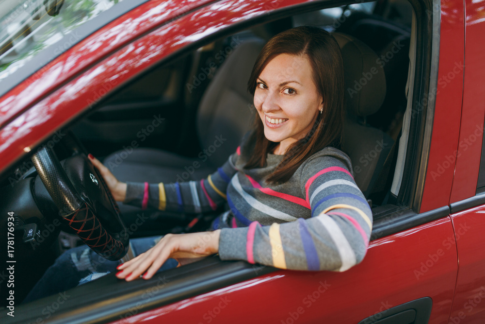 Beautiful young happy smiling European brown-haired woman with healthy clean skin dressed in a striped t-shirt sits in her red car with black interior. Traveling and driving concept.