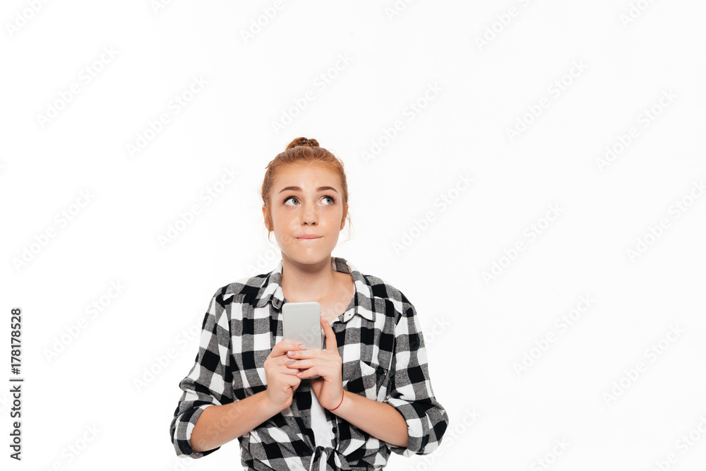 Thoughtful ginger woman in shirt holding smartphone and looking up
