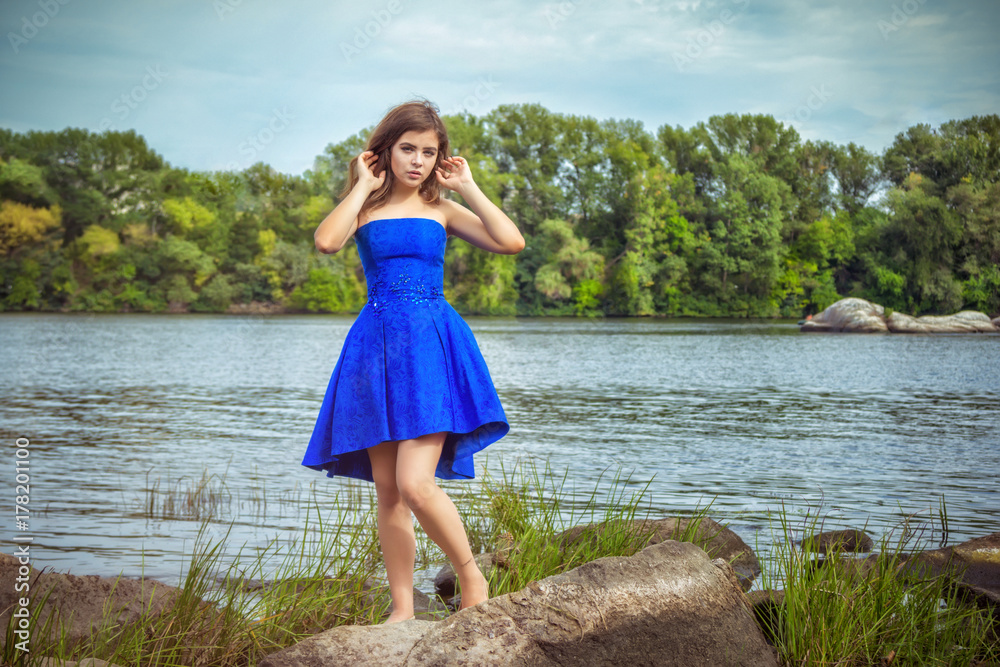Portrait of beautiful young brunette woman, wearing elegant blue dress, standing on a stone river bank with green trees and blue sky on background Portrait of beautiful young brunette woman, wearing elegant blue dress, standing on a stone river bank with green trees and blue sky on background