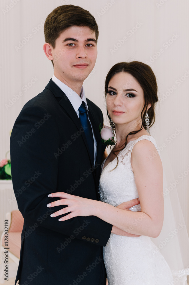 The bride in a white dress and groom in a blue suit are standing in the room and holding a wedding bouquet. Close-up portrait. Happy newlyweds The bride in a white dress and groom in a blue suit are standing in the room and holding a wedding bouquet. Close-up portrait. Happy newlyweds
