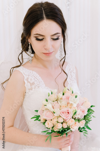 Beautiful bride perfect style. Wedding hairstyle make-up dress and bride's bouquet. Young attractive bride on chair in white room at studio