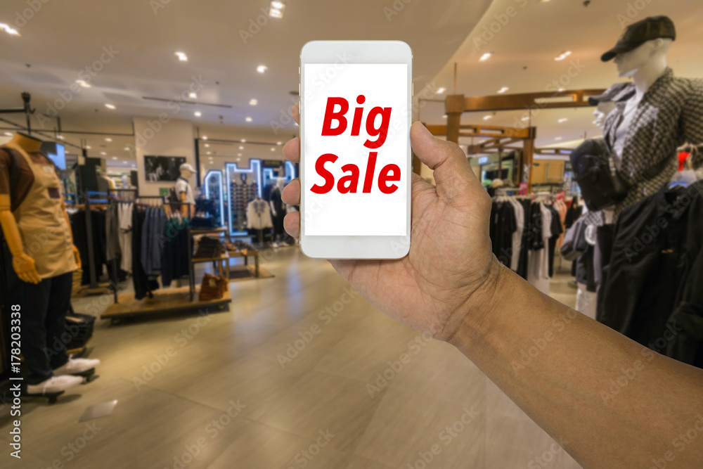 Fototapeta premium Hand holding phone in a clothes store of a shopping mall. Big sale on the screen of the smartphone