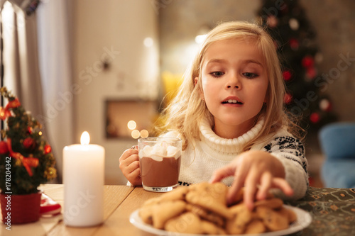 Close-up photo of happy little girl in knitted sweater holding cup of hot chocolate  taking cookie on Christmas morning