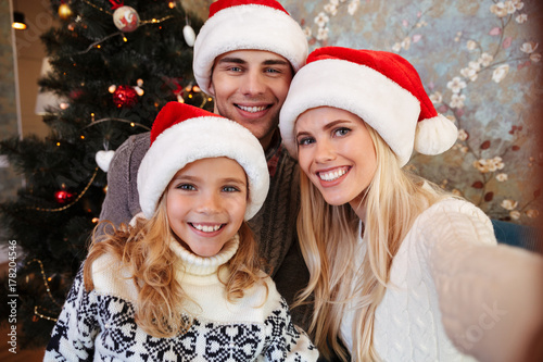 Lovely young family in Santa's hat taking selfie near Christmas tree