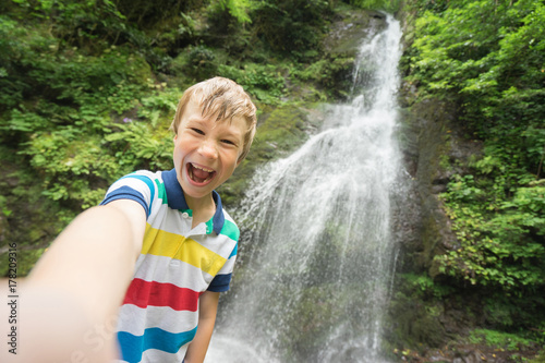 Funny child make selfie with beautiful waterfall on background.